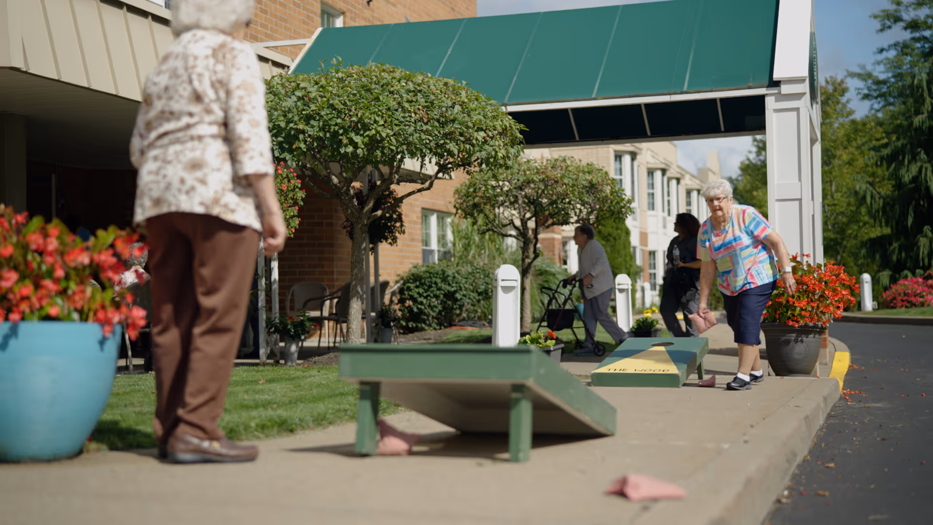 Two elderly women playing cornhole on a sidewalk outside a senior living facility with a green awning and potted flowers, while two other people walk in the background near trimmed trees and a brick building.