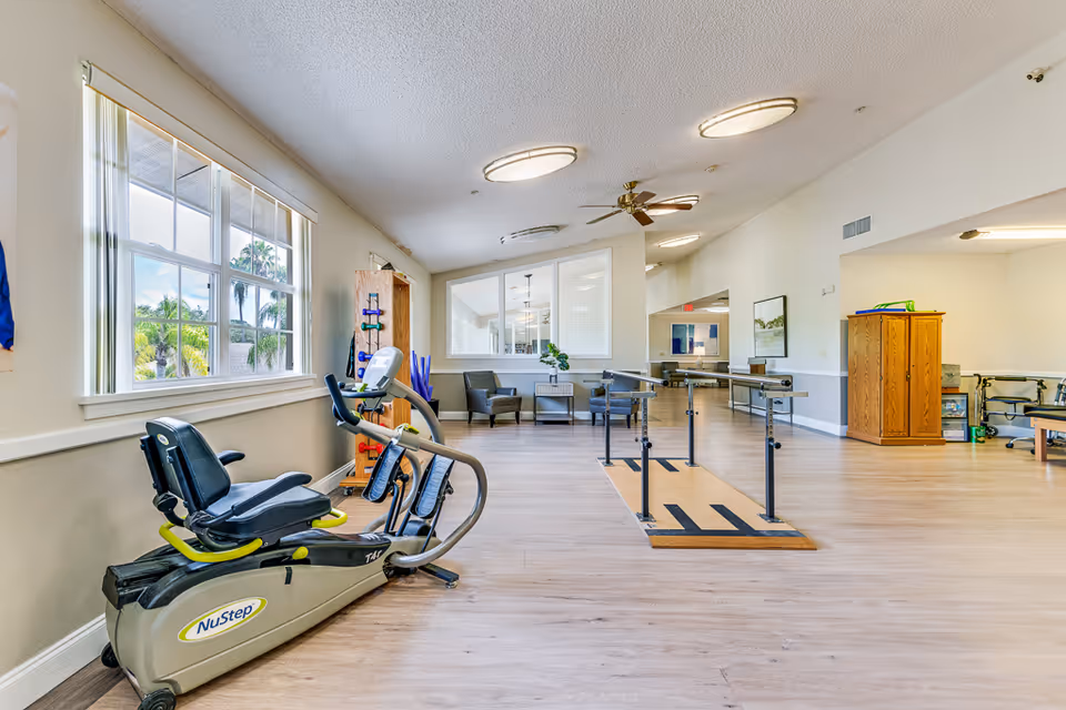 A bright and spacious rehabilitation or exercise room in a senior living facility with a NuStep exercise machine near a large window, parallel bars for walking practice, chairs, and wooden cabinets. The room has light-colored walls and wood flooring.