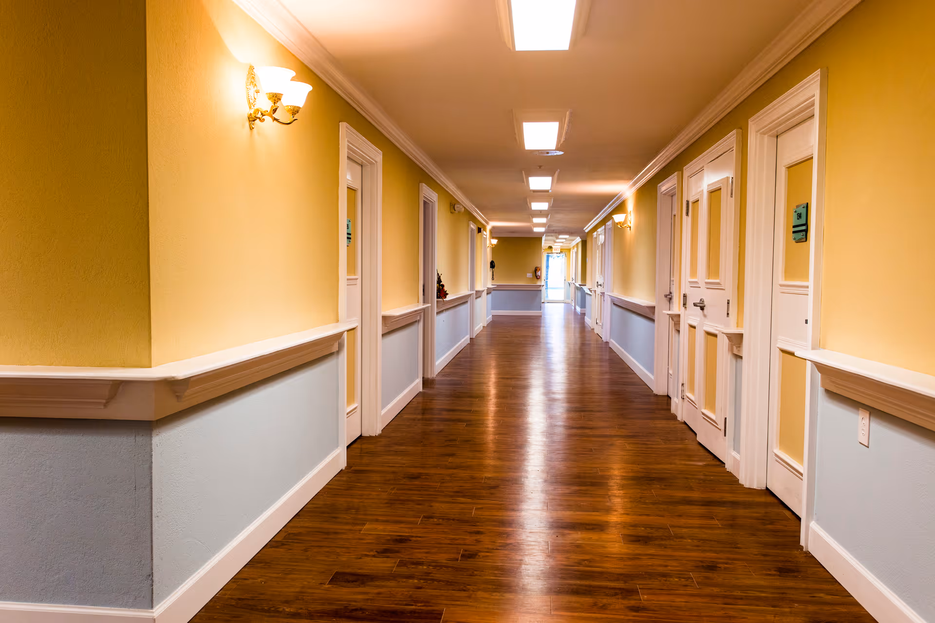 Long, well-lit hallway with wooden floors, yellow walls, and doors along both sides in an assisted living facility.