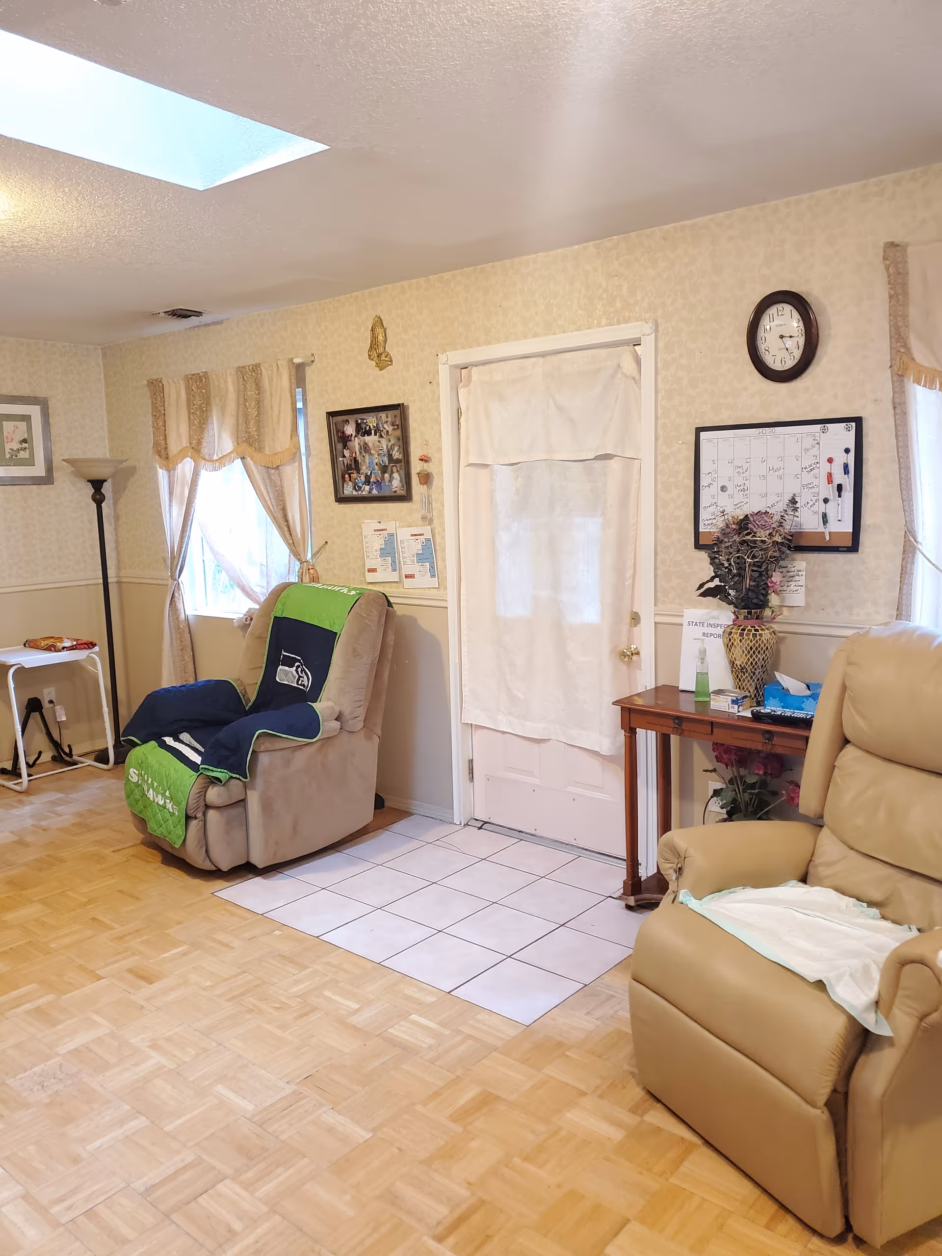A cozy living room area with two recliner chairs, one covered with a green and blue Seahawks blanket. There is a small wooden table with a vase of flowers, tissues, and papers next to a door covered with a white curtain. The walls have light-colored wallpaper with framed pictures, a clock, and a calendar. A floor lamp stands near a window with beige curtains, and the floor is a combination of parquet wood and white tiles near the door.