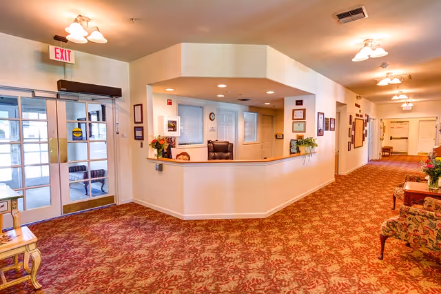 Reception desk and lobby area of a senior living facility with patterned carpet, seating, overhead lights, and an exit door.