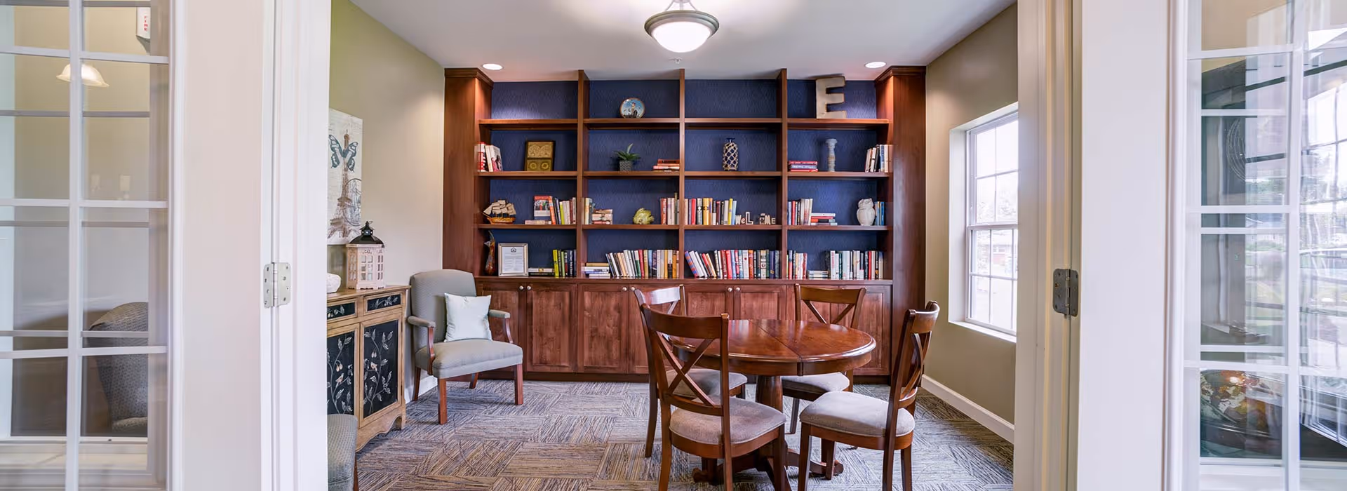 Bright memory-care common room with a built-in wooden bookcase, round wooden table with chairs and an armchair framed by glass doors and windows.
