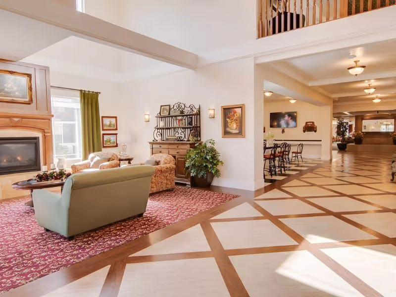 A spacious and well-lit senior living facility common area with a seating arrangement including a green sofa and two patterned armchairs on a red patterned rug near a fireplace. The room features high ceilings, framed artwork on the walls, a decorative wooden cabinet, and a potted plant. The floor has a geometric pattern, and there is a hallway with tables and chairs visible in the background.