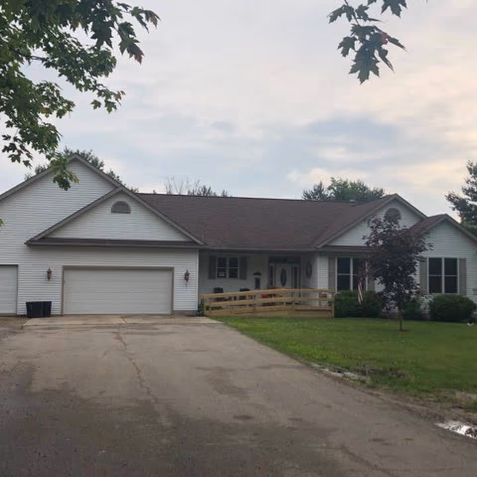 Front exterior view of a single-story white house with a brown roof, a double garage, a wooden ramp leading to the front door, and a green lawn with a small tree on the right side.