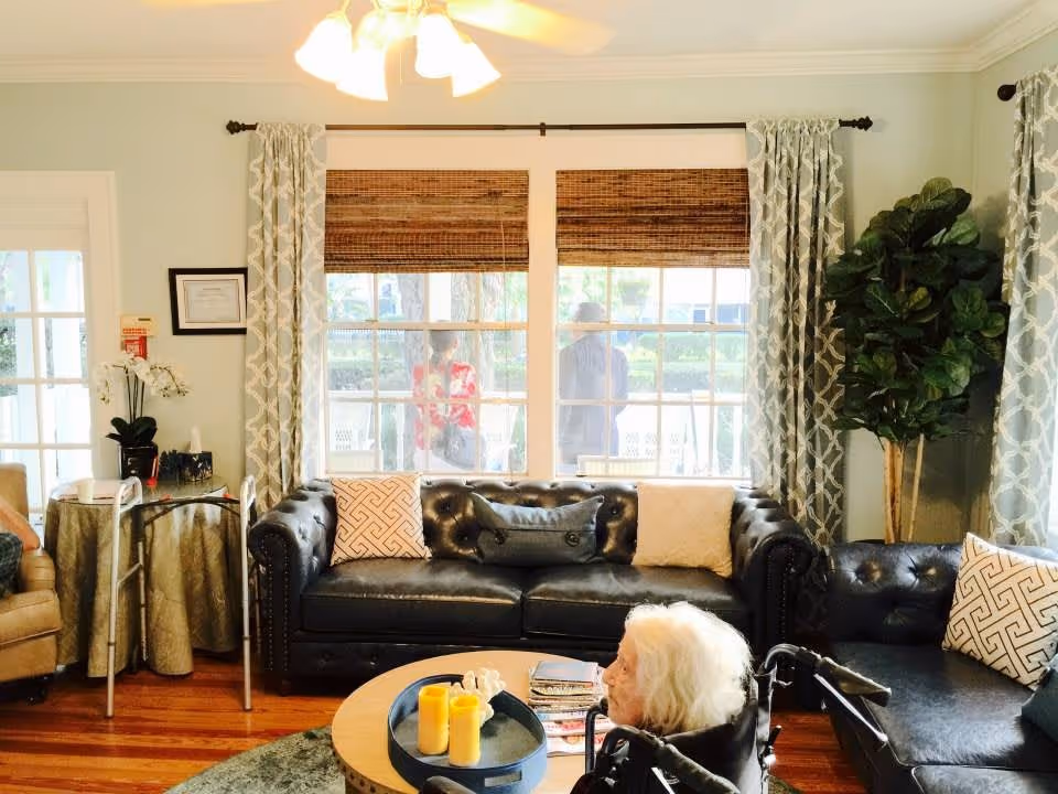 A cozy living room in an assisted living facility with a black leather sofa and armchair, decorative pillows, a round wooden coffee table with candles and magazines, a large potted plant, and a window with bamboo blinds and patterned curtains. An elderly person in a wheelchair is seated near the coffee table, and two people are visible outside through the window.