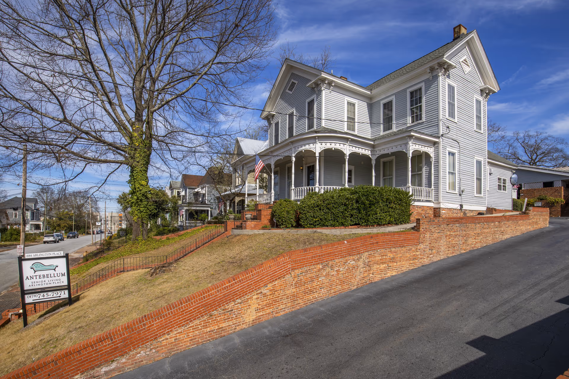 White Victorian-style senior living house with a wraparound porch, American flag, brick retaining walls, and a lawn sign for Antebellum Senior Living.