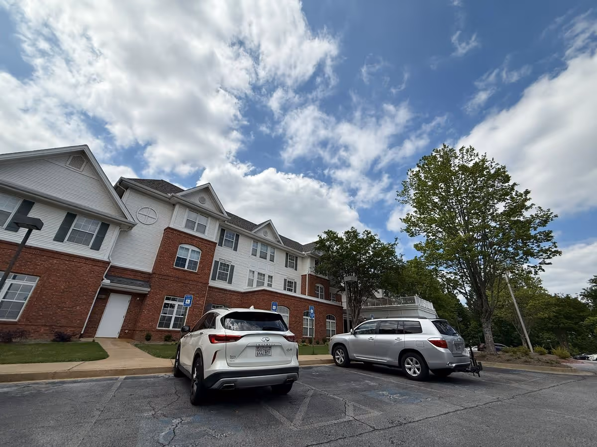Front exterior of a multi-story brick-and-white senior living building with parked cars and trees under a partly cloudy sky.