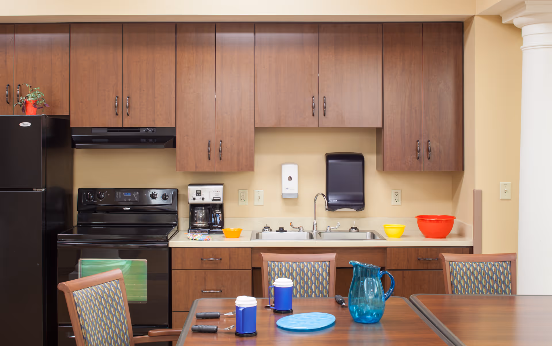 A communal kitchen area with wood cabinets, black refrigerator and stove, a double sink, and a dining table with chairs and pitchers.