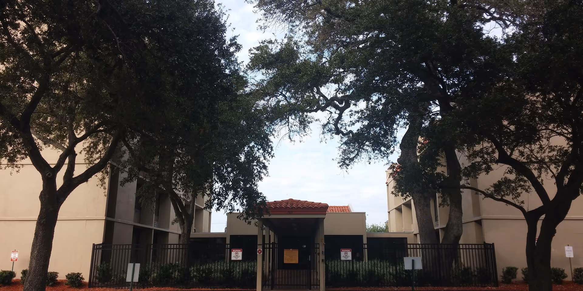 Exterior view of Angels Senior Living at Hacienda Villas showing a gated entrance between two beige buildings with a red-tiled roof over the entrance. Large trees with green foliage frame the entrance area.