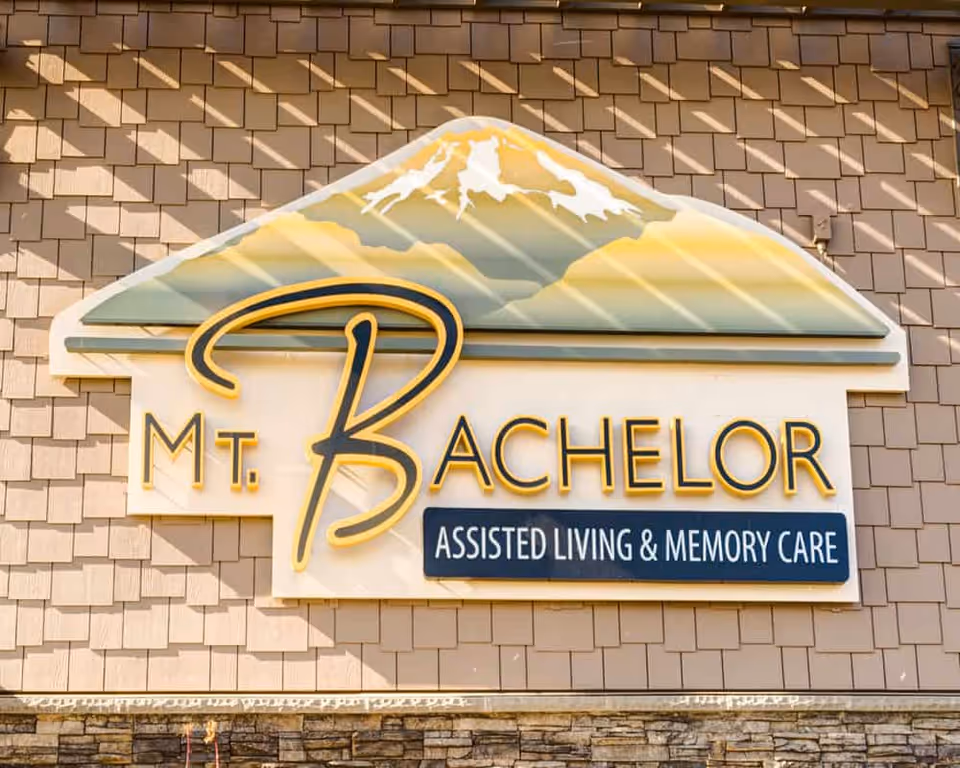 A large decorative sign reading "Mt. Bachelor Assisted Living & Memory Care" with a mountain graphic mounted on a shingled exterior wall.