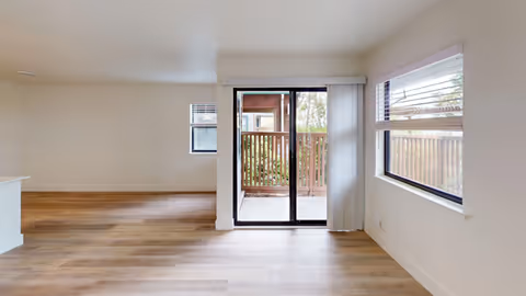 Empty bright living area with wood-look floors, a sliding glass door opening to a small fenced patio and windows on the side walls.