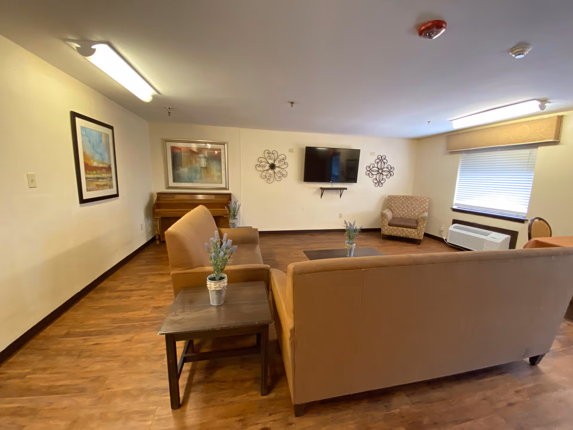 A cozy living room area in a nursing center featuring two brown sofas, a patterned armchair, a wall-mounted flat screen TV, a piano, framed artwork, decorative wall hangings, and a window with blinds. The room has wood flooring and soft lighting from ceiling fixtures.