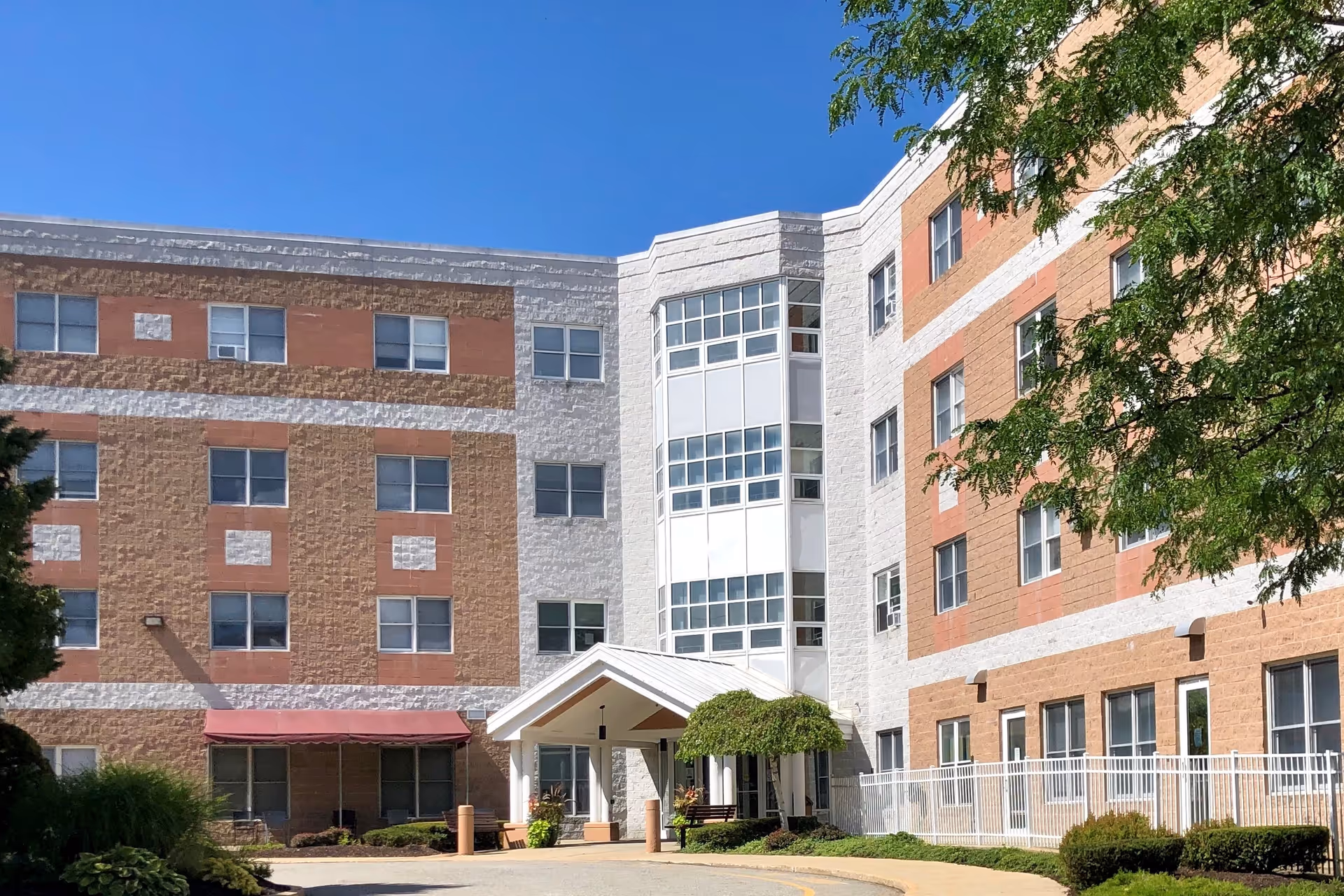 Exterior view of a multi-story senior living facility building with a covered entrance, multiple windows, and landscaping including bushes and trees under a clear blue sky.