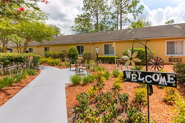 A paved walkway leading through a landscaped garden area with various plants and flowers. There is a decorative metal sign with the word 'WELCOME' in the foreground. The building in the background is a single-story structure with beige walls and several windows. Trees and shrubs surround the area under a partly cloudy sky.