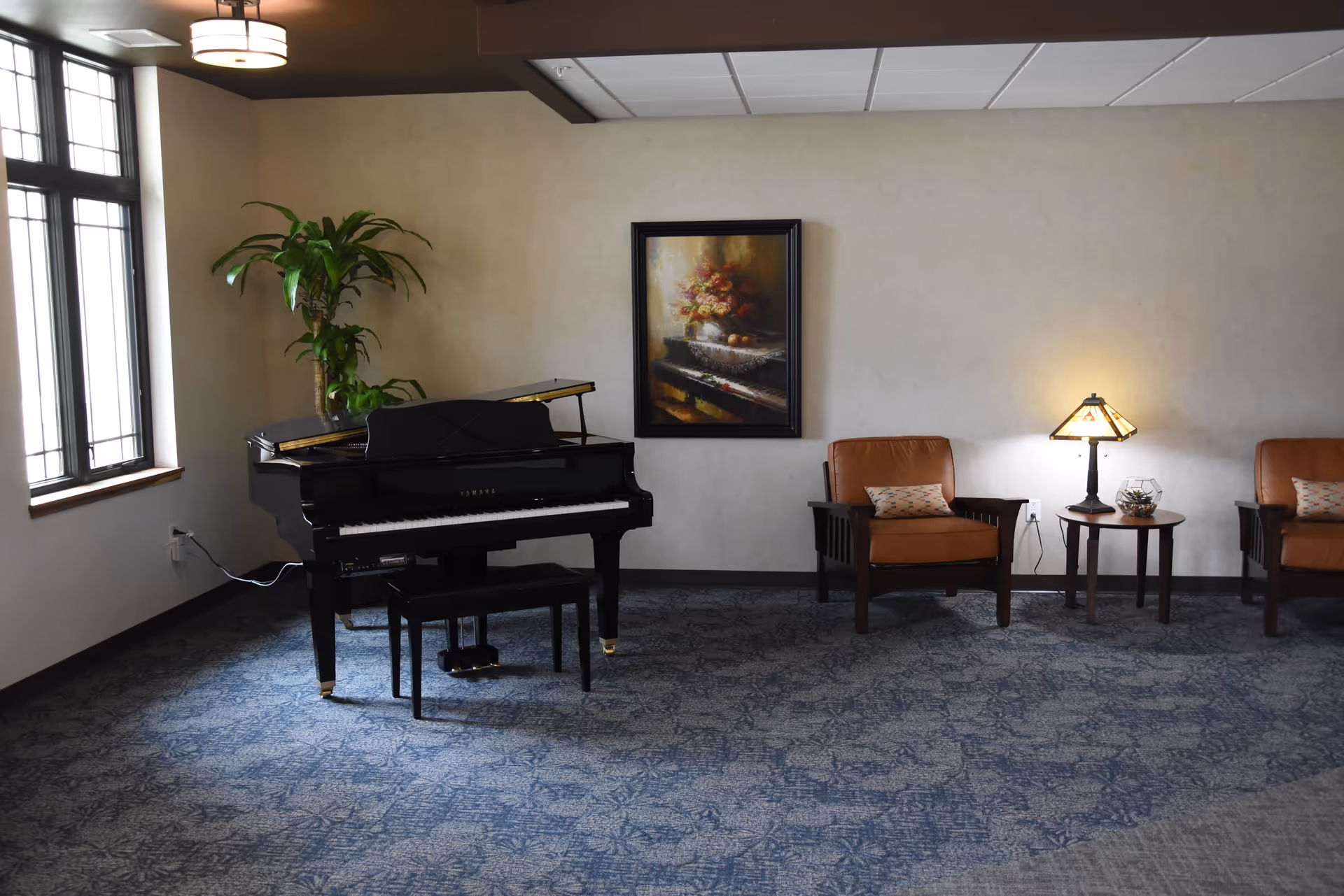 A quiet room with a black grand piano and bench on the left side, a tall green plant behind the piano, two brown armchairs with patterned cushions on the right side, a small round wooden table with a lamp and decorative item between the chairs, and a framed painting of flowers on the wall.