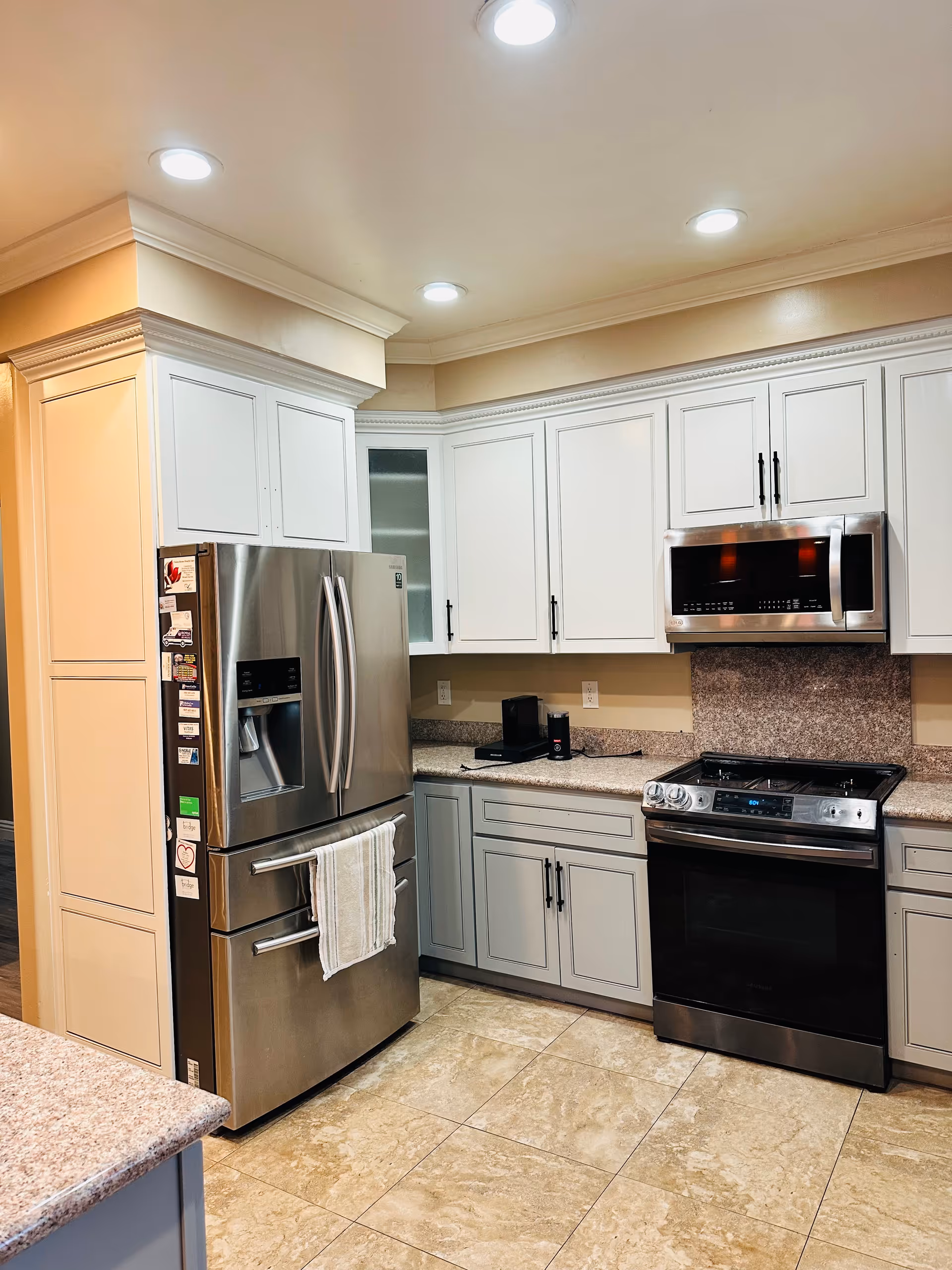 Modern kitchen with white upper cabinets and gray lower cabinets, a stainless steel refrigerator with magnets and a towel hanging on the handle, a stainless steel oven with a stove top, a microwave above the stove, granite countertops, and beige tiled floor.