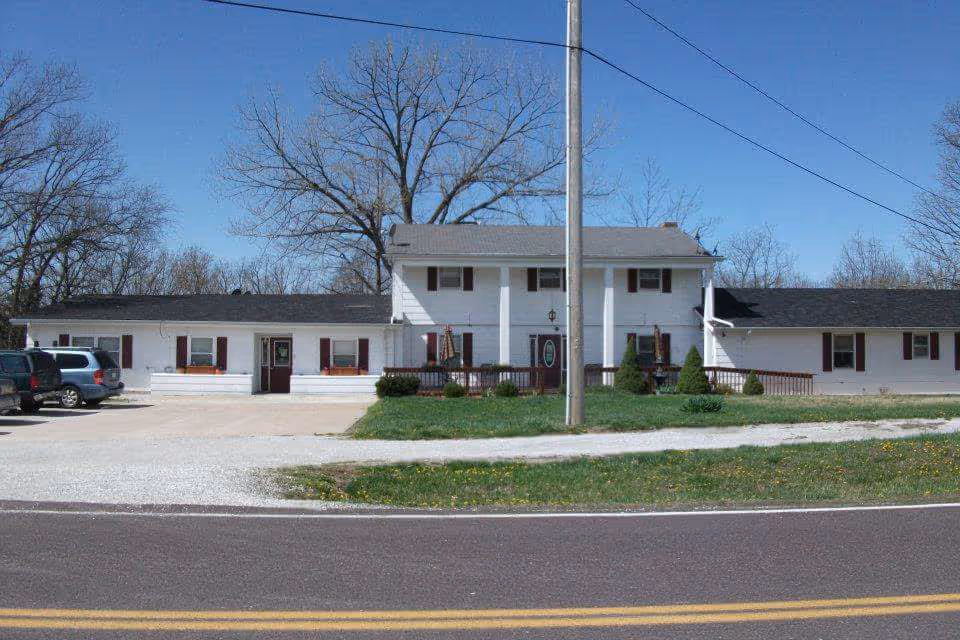 Front exterior of a two-story white building with a driveway, parked cars, lawn, and a utility pole.