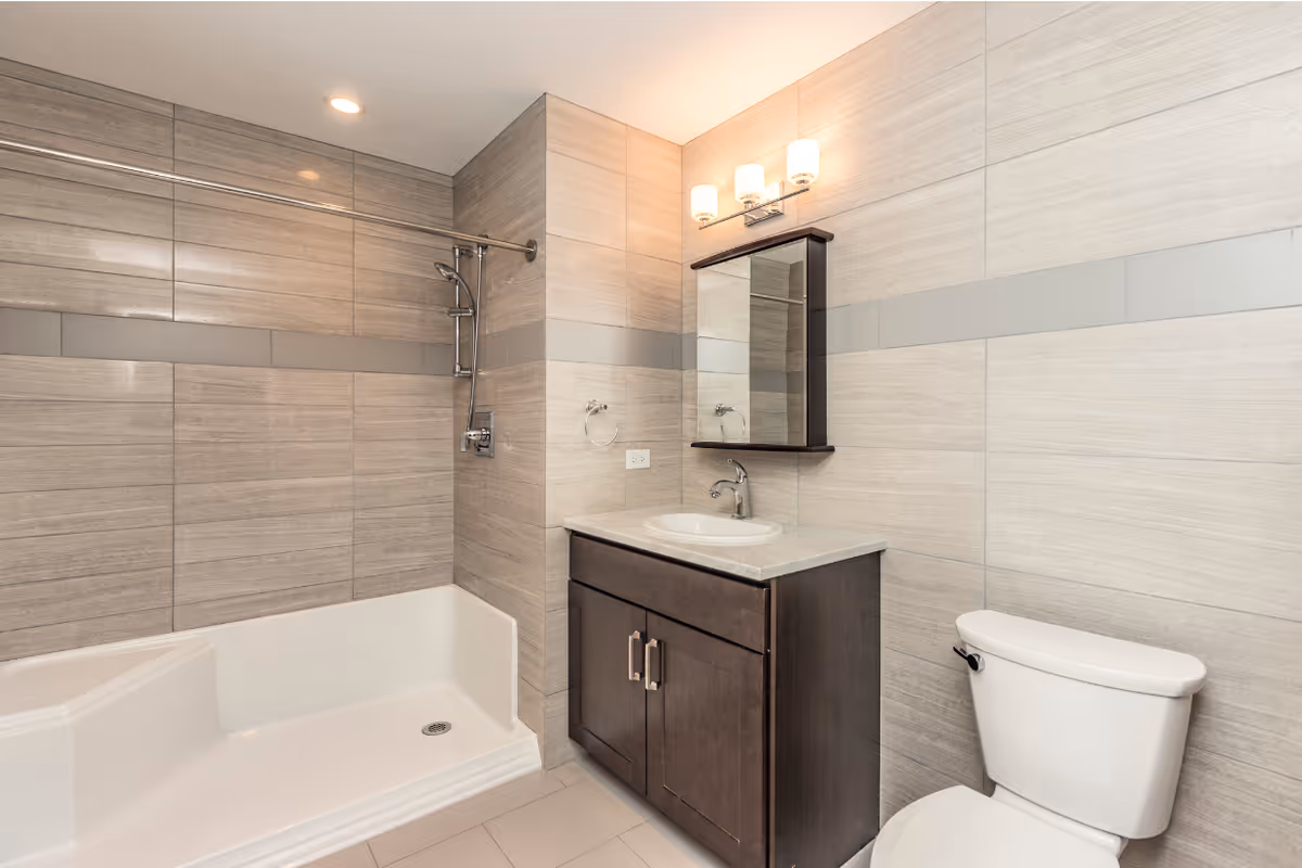 Modern bathroom with light beige tiled walls and floor, featuring a white bathtub with a showerhead, a dark wood vanity with a white sink and marble countertop, a rectangular mirror with a small shelf, and a white toilet.