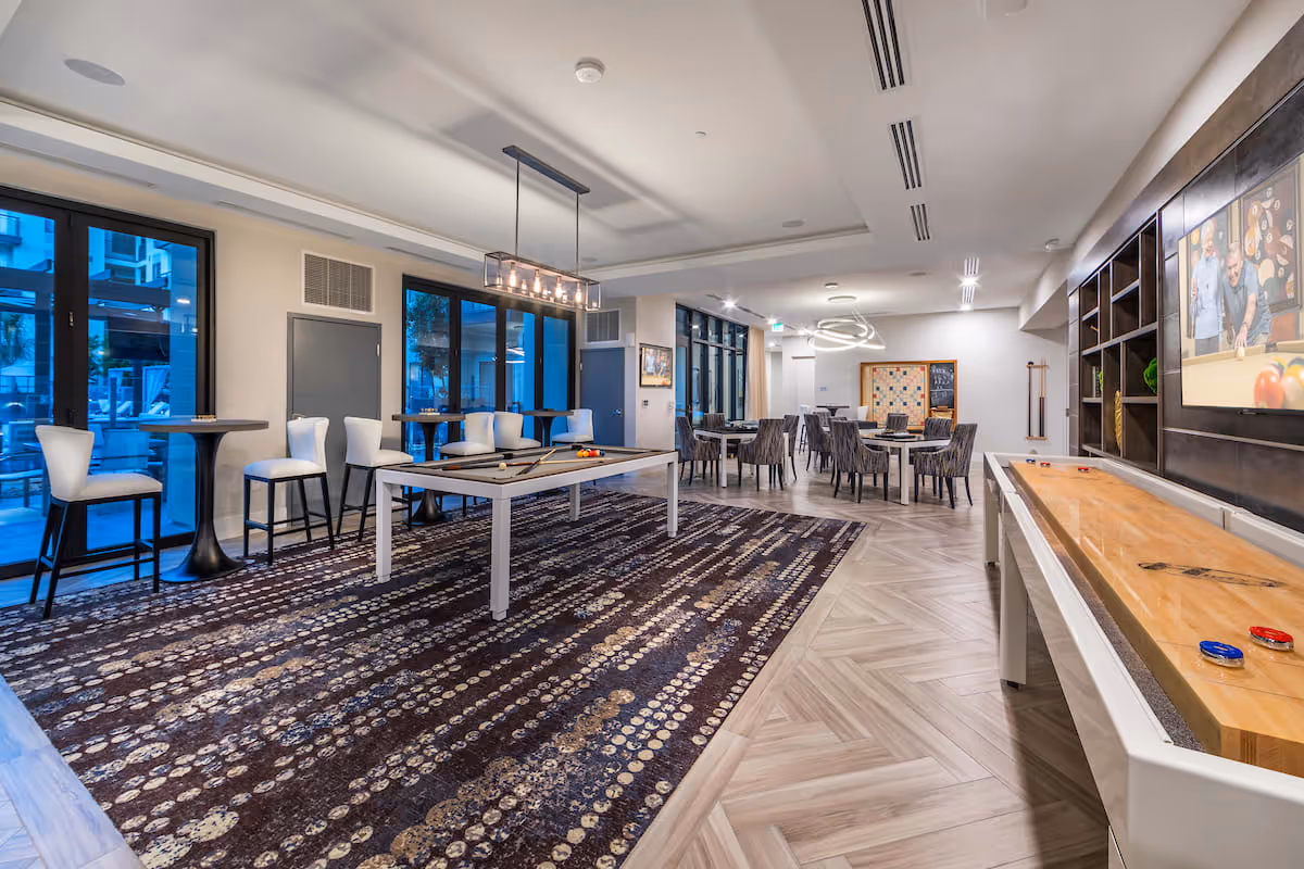 A spacious and modern game room featuring a pool table with balls and cues, a shuffleboard table with pucks, several high tables with chairs near large windows, and multiple dining tables with chairs in the background. The room has a patterned carpet, wood flooring, and contemporary lighting fixtures.