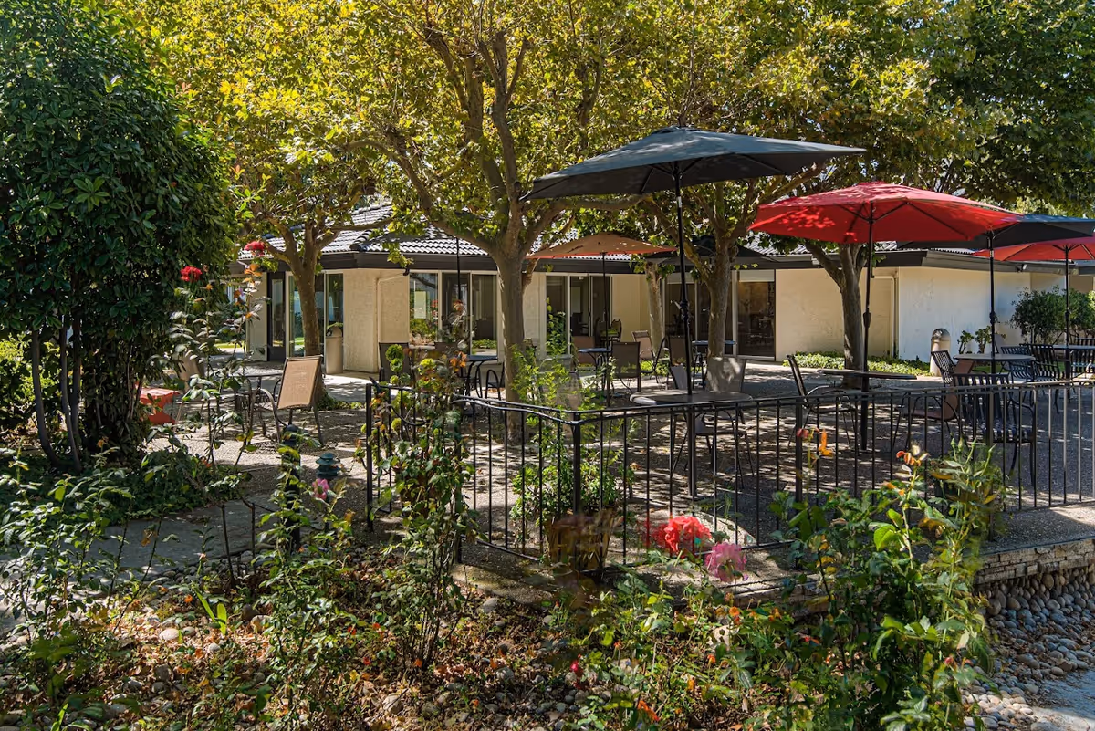 Outdoor patio area at Villa Serena Retirement Community with tables, chairs, and umbrellas under trees surrounded by plants and flowers.