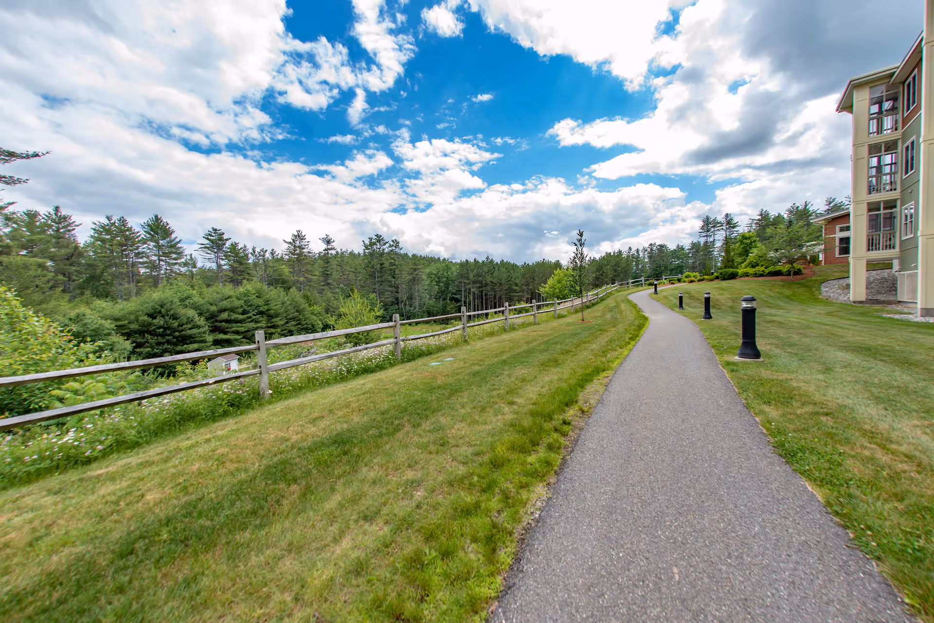 A paved walking path beside a grassy area with black lamp posts leading towards a multi-story building on the right. On the left side, there is a wooden fence with dense green trees and a partly cloudy blue sky above.