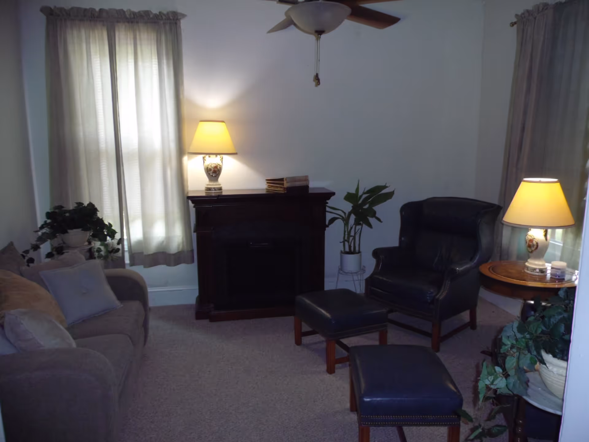 A cozy living room with a beige sofa on the left, a dark leather armchair with matching ottomans in the center, two table lamps on wooden tables, a dark wooden fireplace mantel, and several potted plants. Light beige curtains cover the windows.