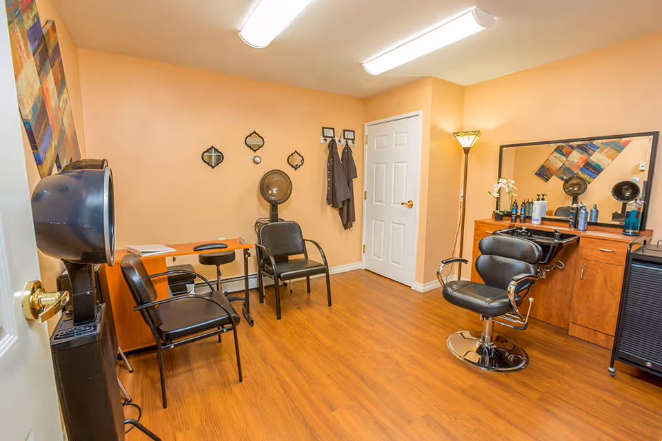 A small hair salon room with peach-colored walls and wooden flooring. The room contains a black salon chair in front of a wooden counter with a large mirror, various hair care products, and a sink. There are two black waiting chairs, a hair dryer chair, a small desk with a stool, and a floor lamp in the corner. Decorative wall mirrors and a colorful abstract painting are also visible.