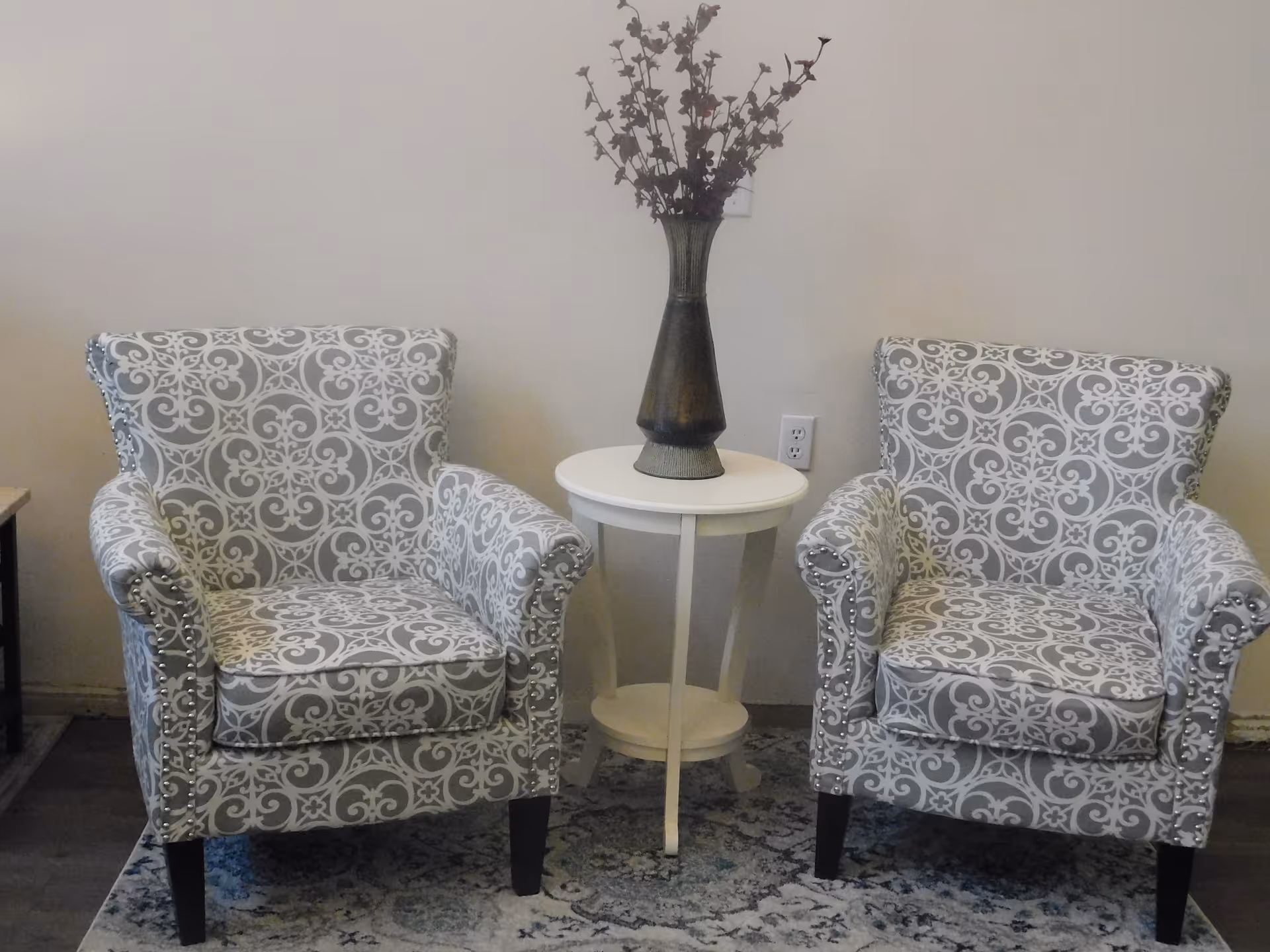 Two patterned armchairs with a white and gray floral design are placed on a patterned rug. Between the chairs is a small white round table with a tall vase holding dried flowers. The background is a plain light-colored wall with an electrical outlet visible.