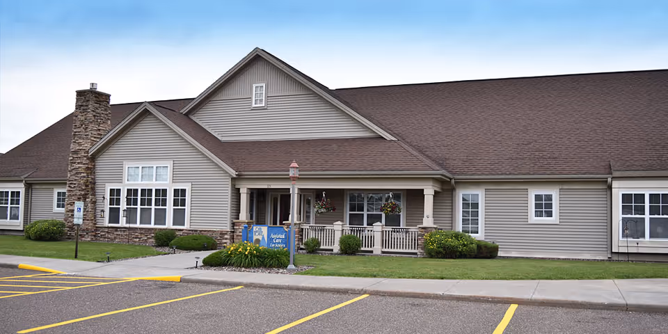 Exterior view of a single-story senior living facility building with beige siding, a brown roof, and a stone chimney. The entrance features a covered porch with hanging flower baskets and a blue sign that reads 'Assisted Care for Seniors.' There is a parking lot with marked spaces in front of the building.
