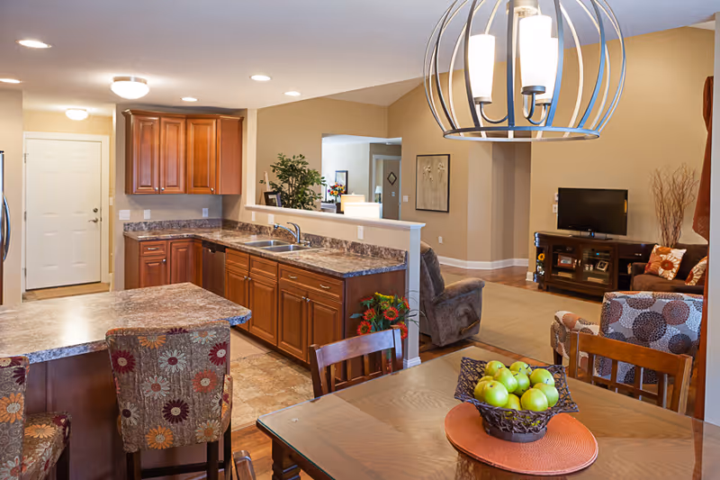 View of a warm-toned open kitchen and living room area in a senior living facility. The kitchen features wooden cabinets, a marble countertop, a sink, and a small breakfast bar with floral-patterned chairs. The dining table in the foreground has a basket of green apples on an orange placemat. The living room has a TV on a wooden stand, a recliner, and patterned armchairs. A modern chandelier hangs above the dining table.