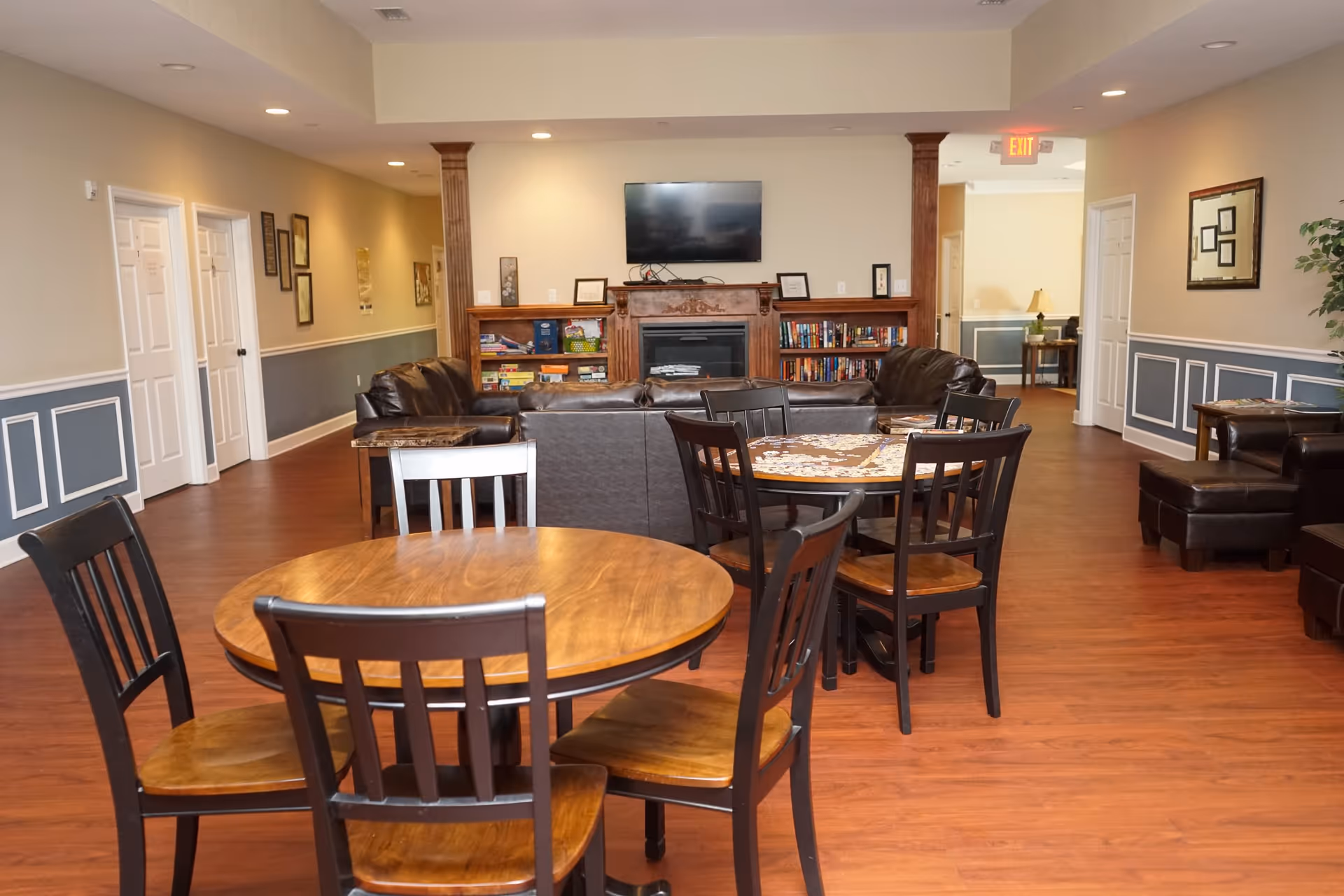 A cozy common area in a senior living facility featuring wooden round tables with chairs, leather sofas arranged around a fireplace with a mounted flat-screen TV above it, and bookshelves filled with books and games. The room has wood flooring, beige walls with blue wainscoting, and framed artwork on the walls.