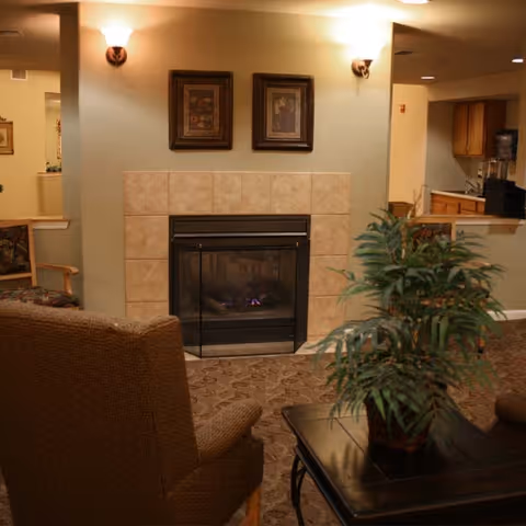 Cozy living room area with a tiled fireplace in the center, two framed pictures above it, wall sconces on either side, a patterned armchair facing the fireplace, a coffee table with a potted plant, and a partial view of a kitchen area in the background.