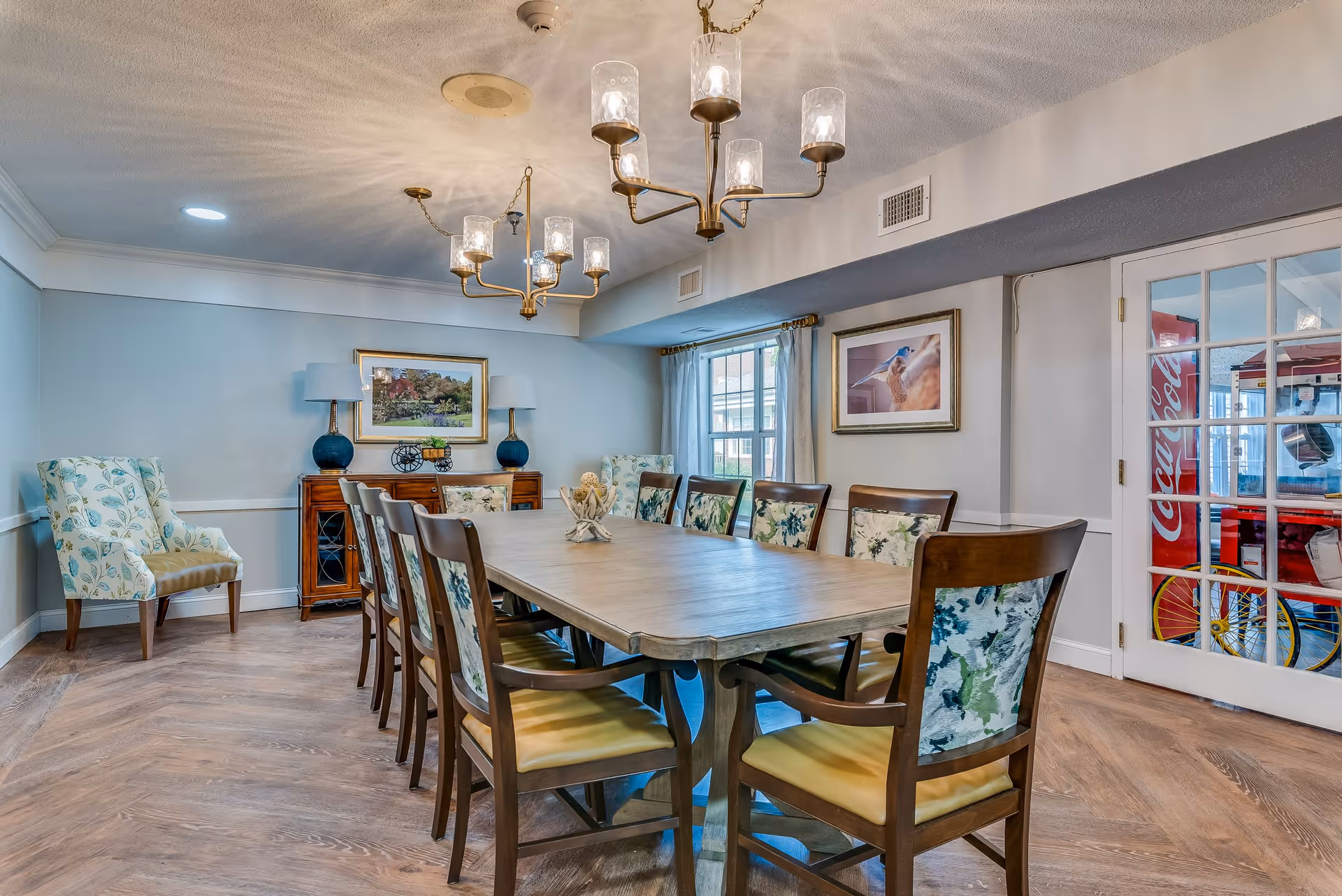 A well-lit dining room with a long wooden table surrounded by ten chairs with floral upholstery. Two chandeliers with glass shades hang from the ceiling. The room has light blue walls, wooden flooring, and two framed pictures on the walls. There are two upholstered armchairs in the corner and a wooden sideboard with two lamps. A glass door on the right reveals a Coca-Cola vending machine in an adjacent room.