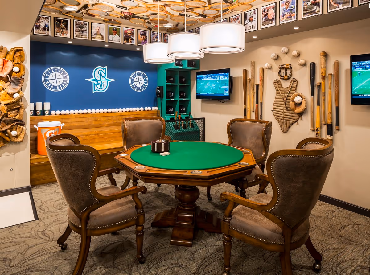 A cozy game room with a green felt poker table surrounded by four brown leather chairs. The walls are decorated with baseball memorabilia including bats, gloves, balls, and a catcher's chest protector. Two flat-screen TVs are mounted on the walls showing a football game. Above the wooden bench seating, there are Seattle Mariners logos and framed photographs of baseball players lining the top of the walls.