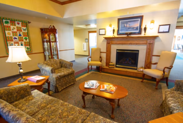 Cozy seating area with sofas, armchairs, a coffee table, and a fireplace in a senior living facility lounge.