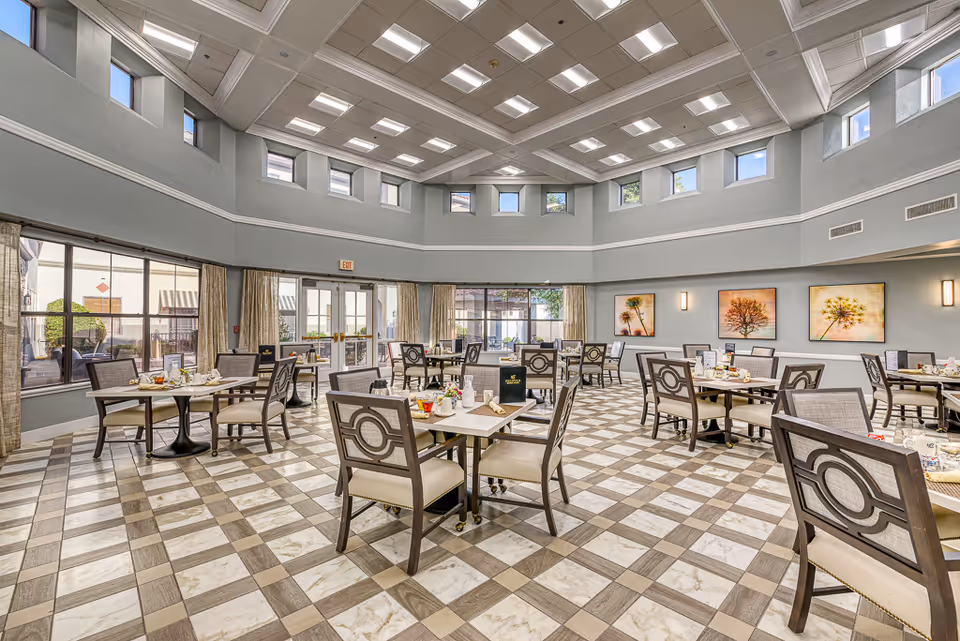A spacious dining room with multiple tables and chairs arranged neatly. The room features a high ceiling with numerous recessed lights and small windows near the ceiling. Large windows and glass doors allow natural light to enter, and the walls are decorated with framed artwork of trees and flowers. The floor has a checkered pattern with light and dark tiles.
