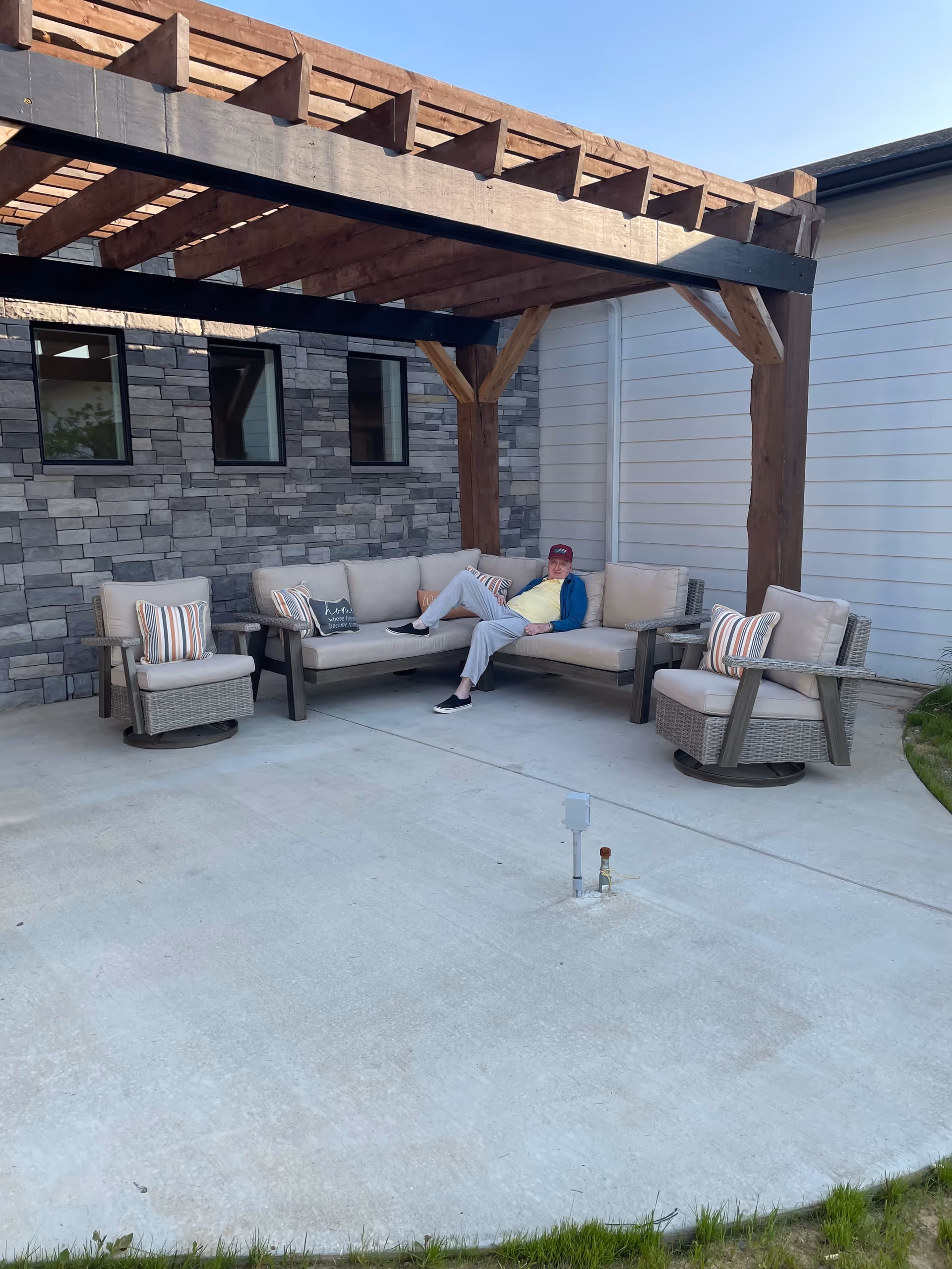 An elderly man wearing a red cap, yellow shirt, blue jacket, and light gray pants is sitting on a beige outdoor sectional sofa under a wooden pergola. The seating area includes two cushioned armchairs with striped pillows. The background features a stone wall with three windows and a white siding wall. The ground is a concrete patio with some grass visible at the edge.