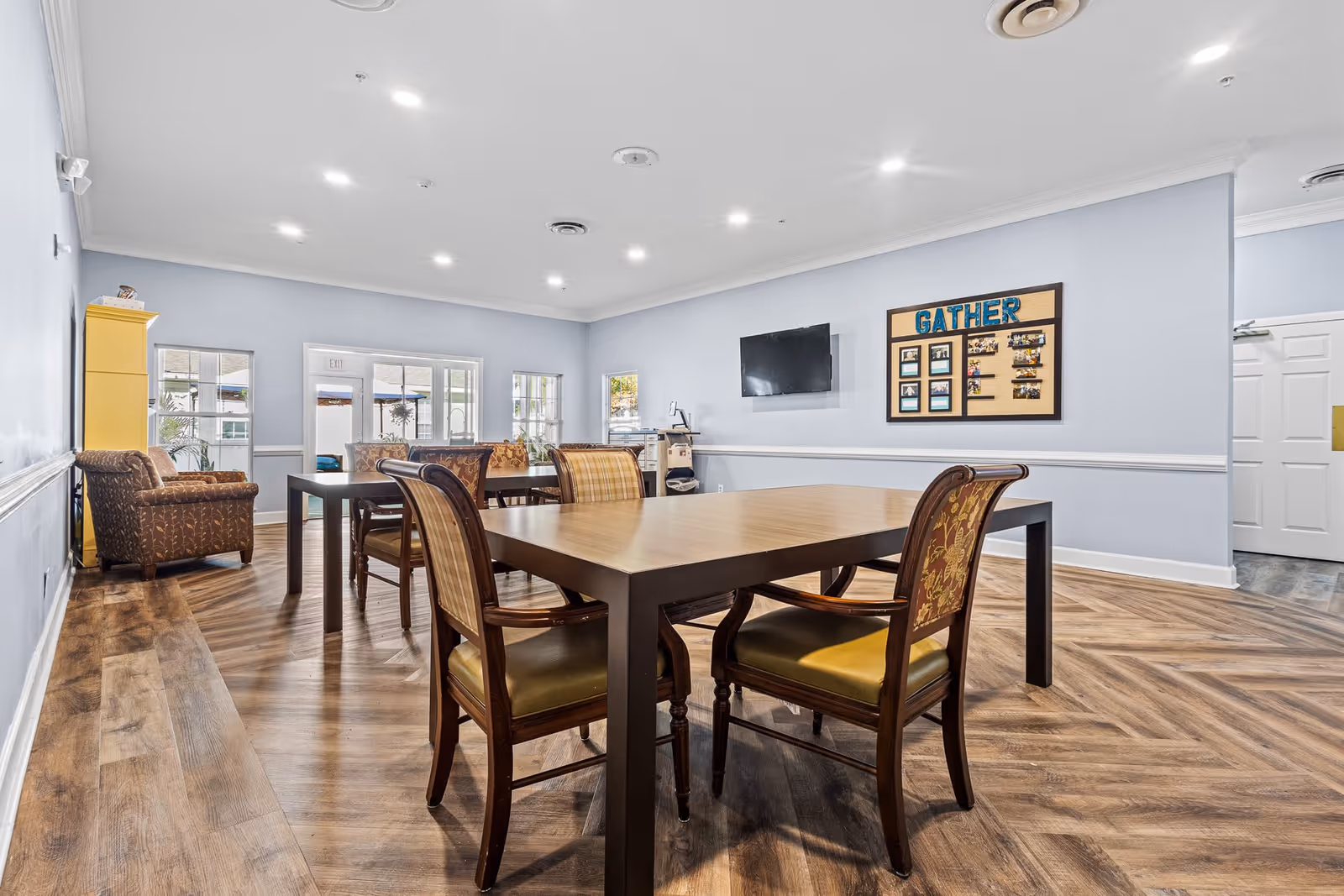 A spacious, well-lit common area with wooden flooring and light blue walls. The room features a large wooden table surrounded by chairs with patterned upholstery. In the background, there are additional chairs, a yellow cabinet, windows, a wall-mounted TV, and a bulletin board with the word 'GATHER' displayed prominently.