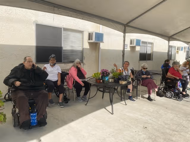 A group of elderly people sitting outdoors under a canopy next to a building. Some are seated in wheelchairs and others in chairs around a table with potted plants. The building wall has windows with air conditioning units.