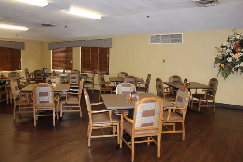 A dining room with multiple square tables and upholstered chairs on a wood floor and a floral arrangement on the wall.