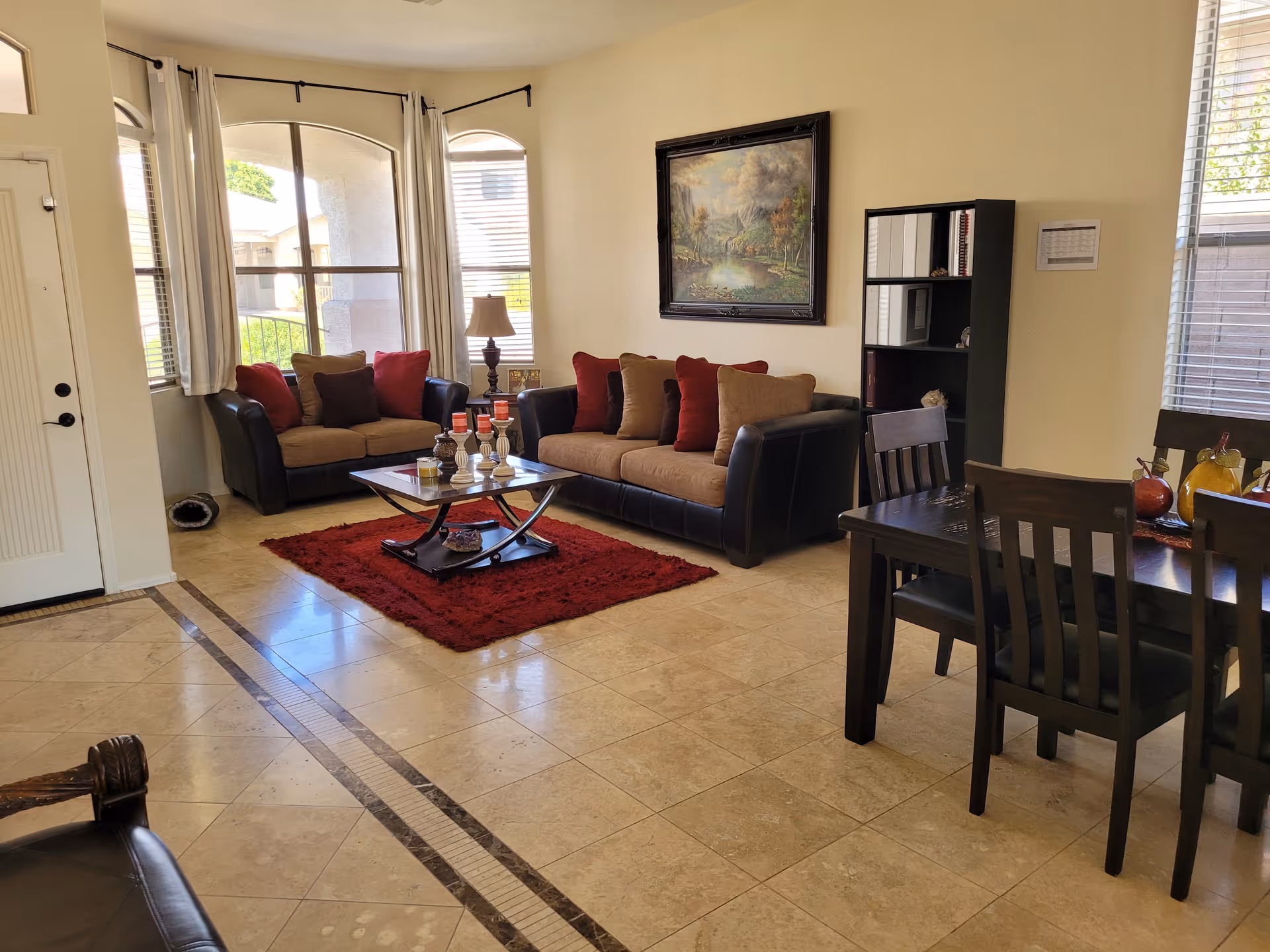 Sunlit living room with two sofas, a coffee table on a red rug, a bookshelf, and an adjacent dining table.