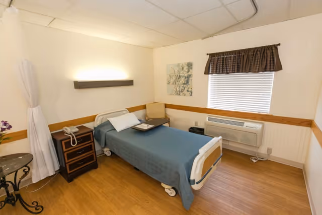 A simple and clean patient room with a single hospital bed covered with a blue blanket, a bedside table with a telephone, a small round table with a flower vase, a chair, a window with blinds and a brown valance, and a wall-mounted air conditioning unit.