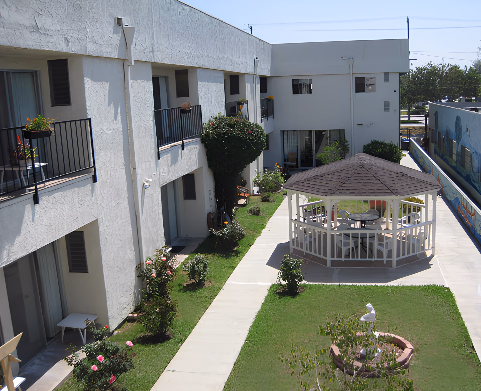 Outdoor courtyard area of a retirement center featuring a white gazebo with chairs and tables inside, surrounded by a walkway and green grass with flower beds. The courtyard is bordered by a two-story white building with balconies and windows, and a mural painted on a wall to the right.