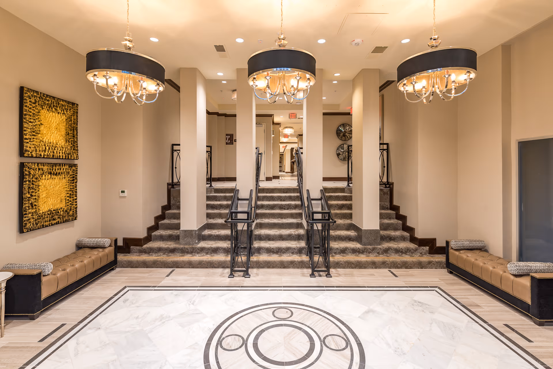 Interior view of a senior living facility lobby area with marble flooring featuring a circular pattern, two brown cushioned benches with patterned pillows on either side, two large golden abstract wall art pieces on the left wall, and three elegant chandeliers hanging from the ceiling. There are carpeted stairs with black metal railings leading to a hallway in the background.