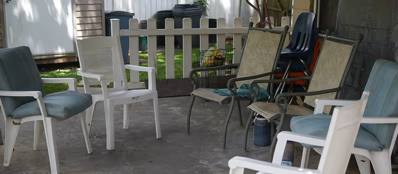 A covered outdoor seating area with a variety of chairs arranged in a circle on a concrete floor. There are cushioned chairs, plastic chairs, and metal chairs. In the background, there is a white picket fence and some outdoor items like trash bins and stacked chairs.