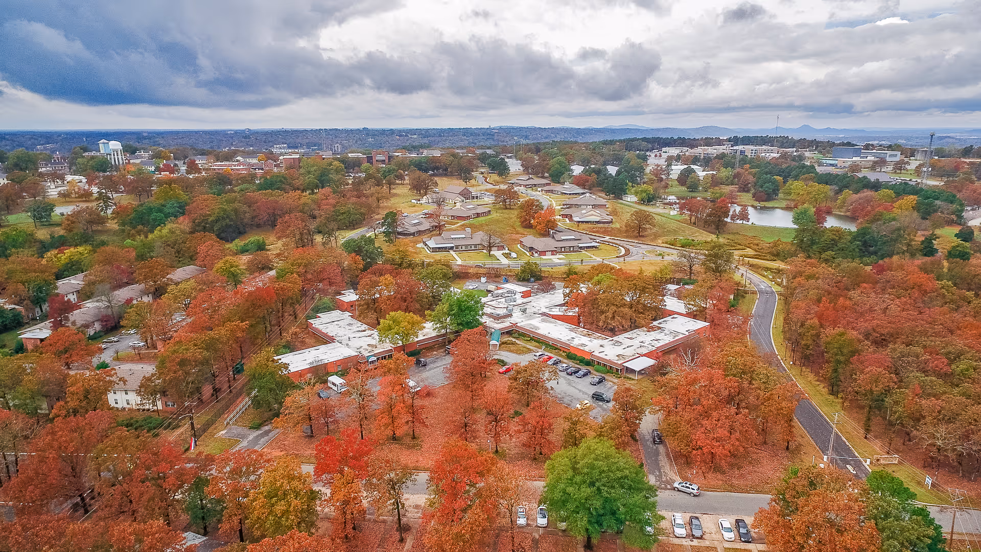 Aerial view of The Blossoms at North Little Rock Rehab & Nursing Center surrounded by trees with autumn foliage. The facility buildings are visible with parking lots and roads winding through the area. The background shows a distant cityscape under a cloudy sky.