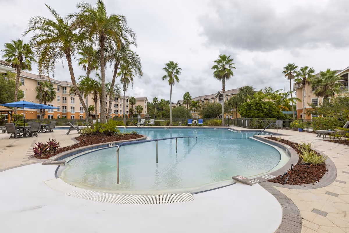Outdoor swimming pool area with palm trees and lounge chairs surrounded by multi-story residential buildings under a cloudy sky.
