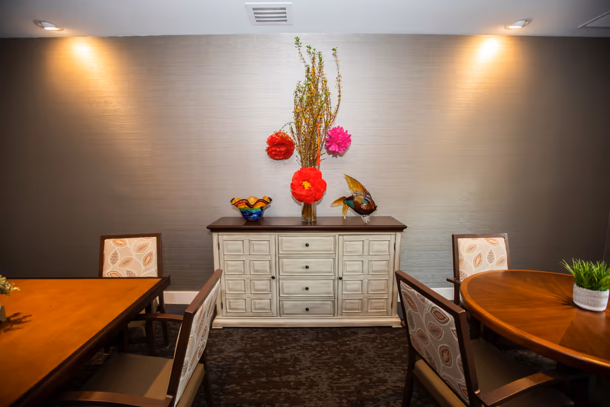 Dining room with wooden tables and upholstered chairs and a white sideboard topped with a vase of flowers and decorative glass pieces.