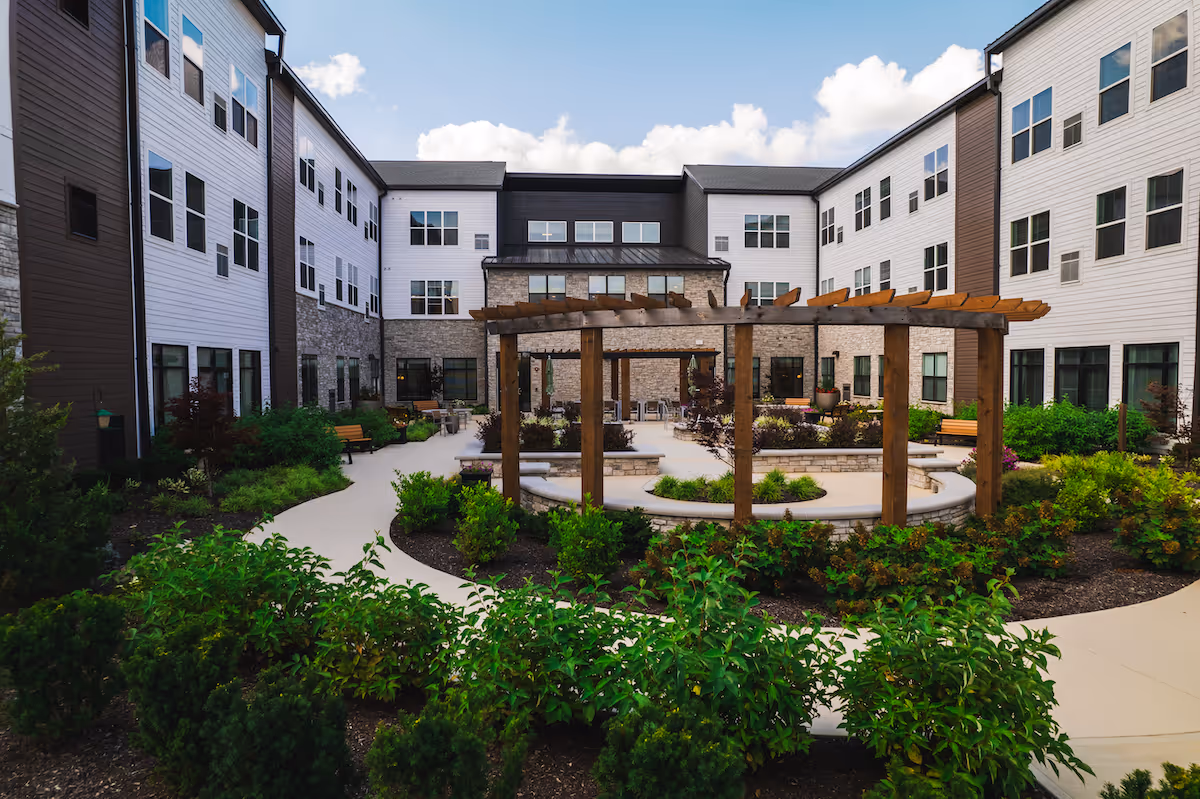 Outdoor courtyard area of a senior living facility with a wooden pergola, curved concrete benches, landscaped garden beds with green shrubs, and a three-story building surrounding the courtyard under a partly cloudy sky.