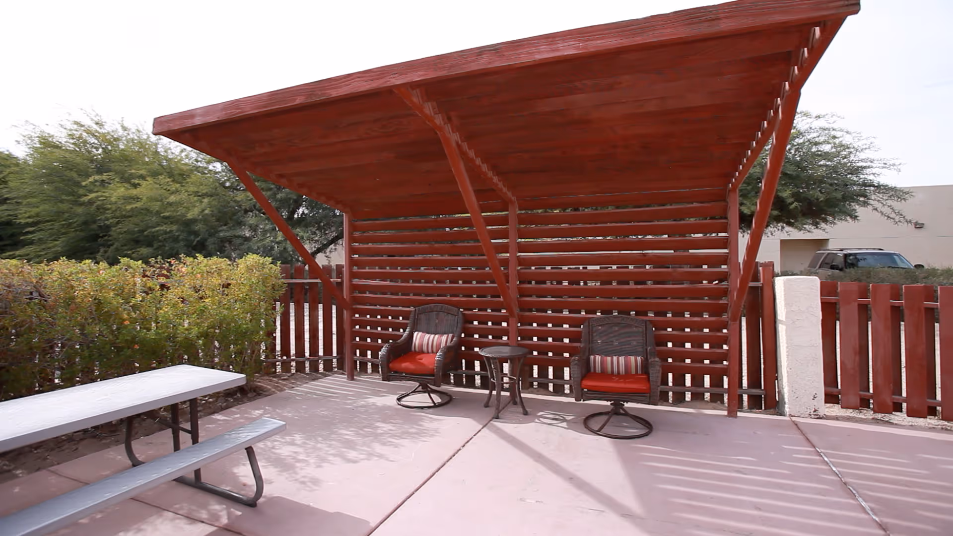 Outdoor patio area with a red wooden pergola providing shade over two cushioned chairs and a small round table. There is a picnic table on the left side and greenery along the fence in the background.